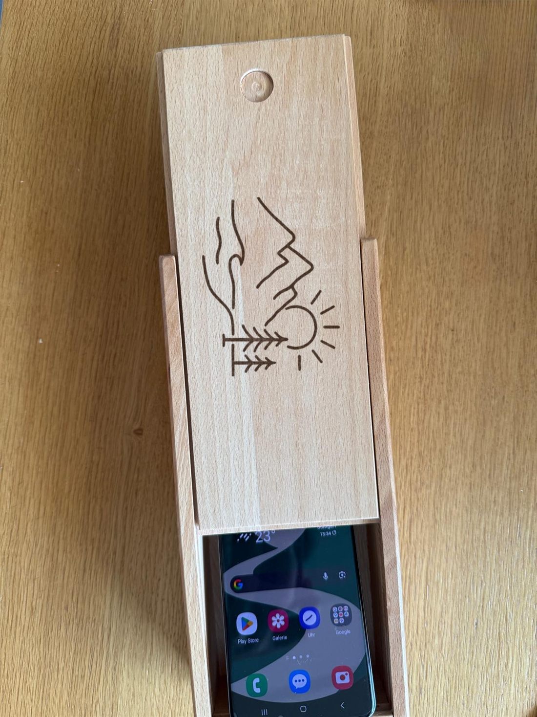Handygarage aus hellem Holz mit eingravierter Zeichnung aus Bergen, Baum und Sonne, davor ein eingeschobenes Smartphone